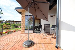 Um pátio externo com chão de tijolos, uma mesa e cadeiras, e um grande guarda-sol.