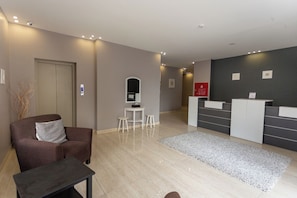 Un espace d’accueil moderne comprenant un comptoir de réception, un coin salon avec un canapé et une petite table, ainsi qu’un miroir au mur.