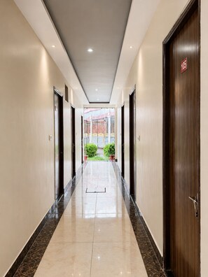 A hotel corridor with a wooden door marked '105' on the right, a glass elevator on the left, and a view of a building with a red roof in the distance. - Madhuvan Resort (Kanpur)