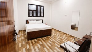 A bedroom with a bed, a desk, a chair, a mirror, and a window with blinds. - Madhuvan Resort (Kanpur)