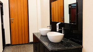 A modern bathroom with a black marble vanity, two white vessel sinks, and a wooden door. - Madhuvan Resort (Kanpur)