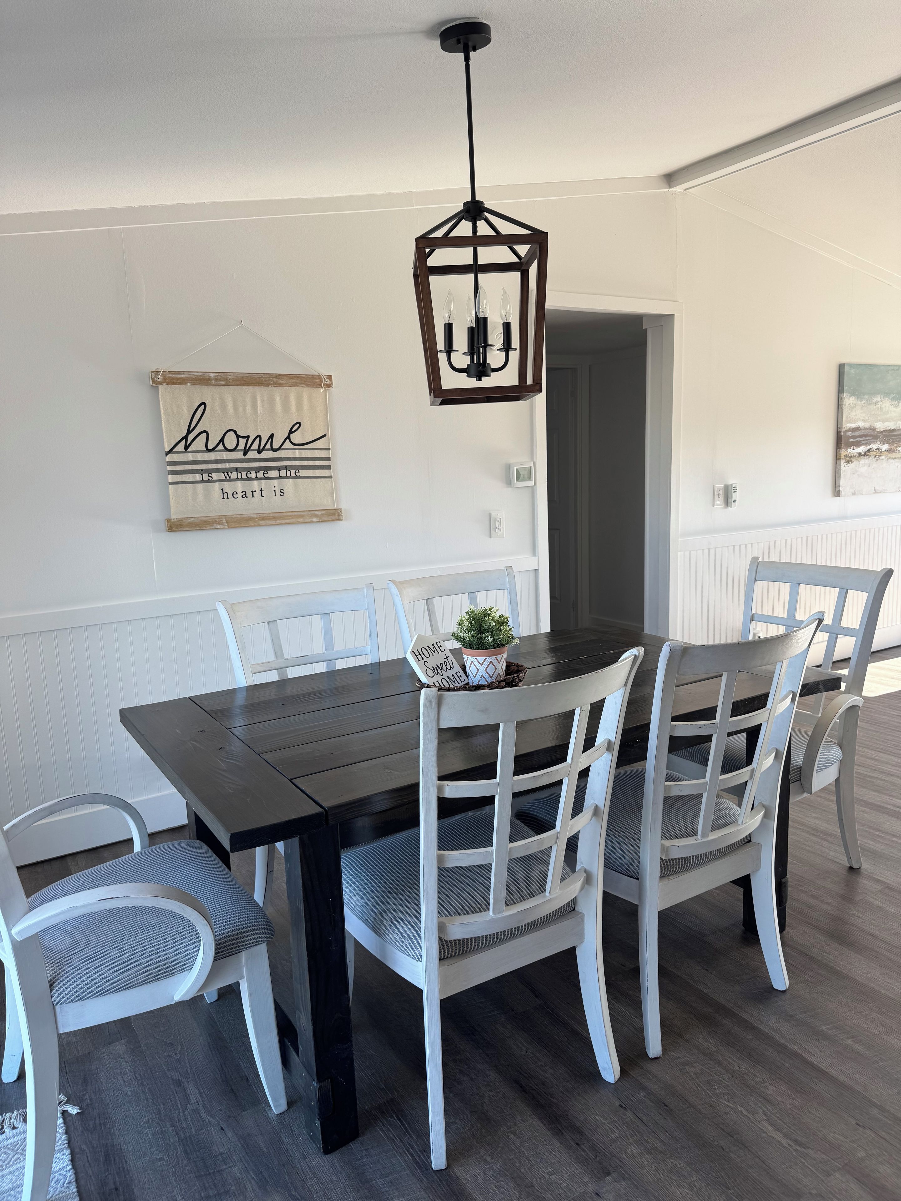 Un comedor con una mesa de madera oscura, sillas blancas, una lámpara colgante y un letrero en la pared.