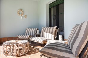 Eine sonnendurchflutete Terrasse mit gestreiften Sofas, einem gewebten Hocker und einem kleinen Tisch mit einer Dekoration.