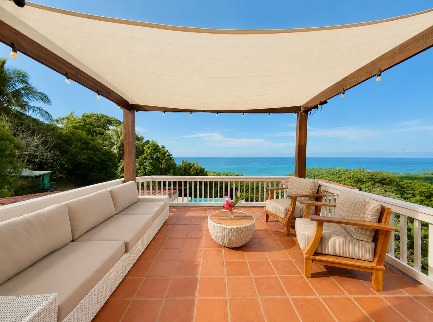 A covered outdoor seating area with a sofa, chairs, and a round table, overlooking a pool and ocean view.