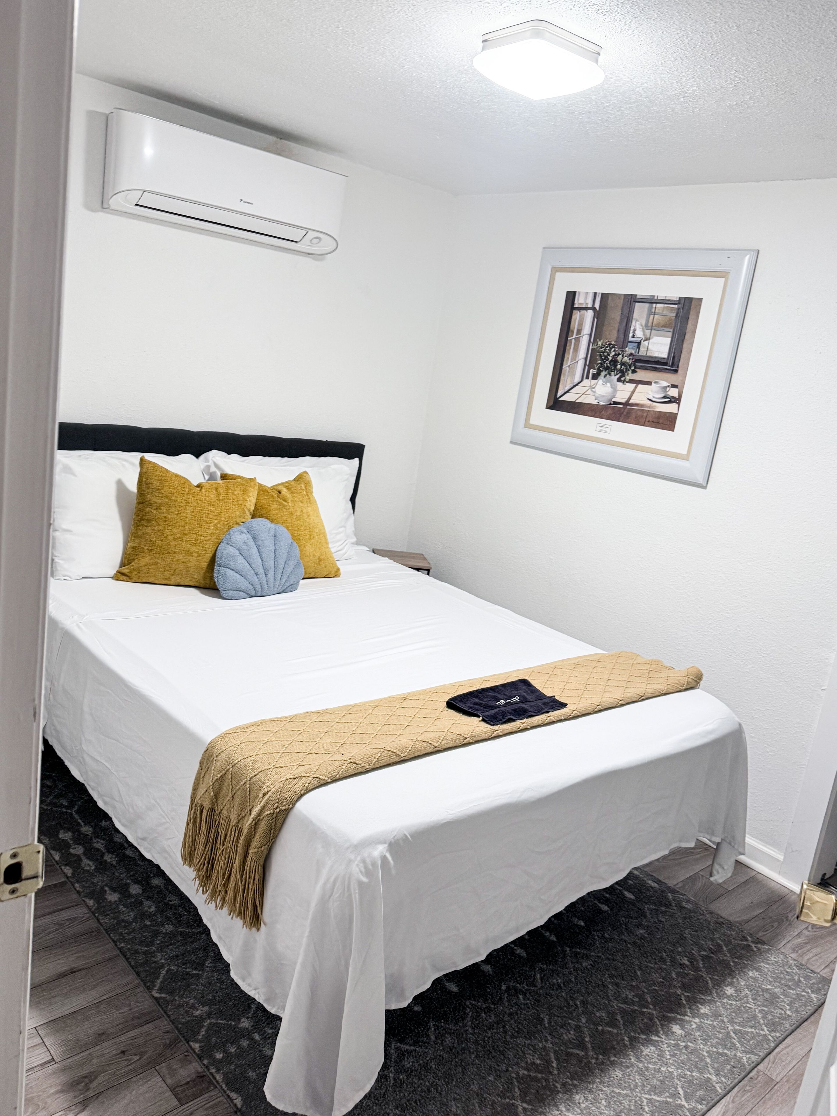 A hotel room with a bed, a bedside table, a wall-mounted air conditioner, a framed picture, and a dark patterned rug.