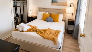 Una cama bien hecha con cabecera, una mesita de noche con lámpara, y una estantería con plantas.
