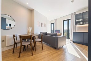 A modern living room with a dining area, a grey sofa, a round table with chairs, and a large window with curtains. - Gym | Coworking | City Centre 2BR | StayCozy (Birmingham)