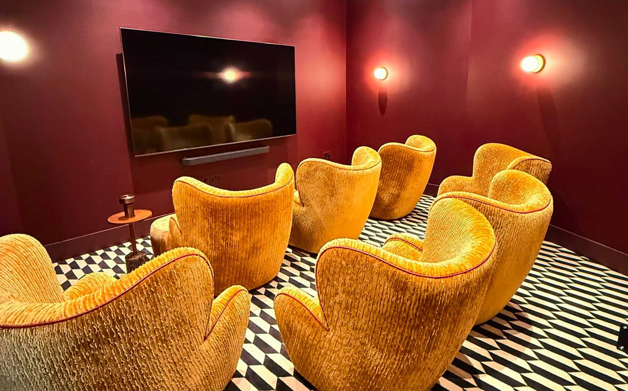 Een kamer met een grote flatscreen televisie aan de muur, verschillende stoelen in een uniek geel design en een vloer met een geruit patroon.