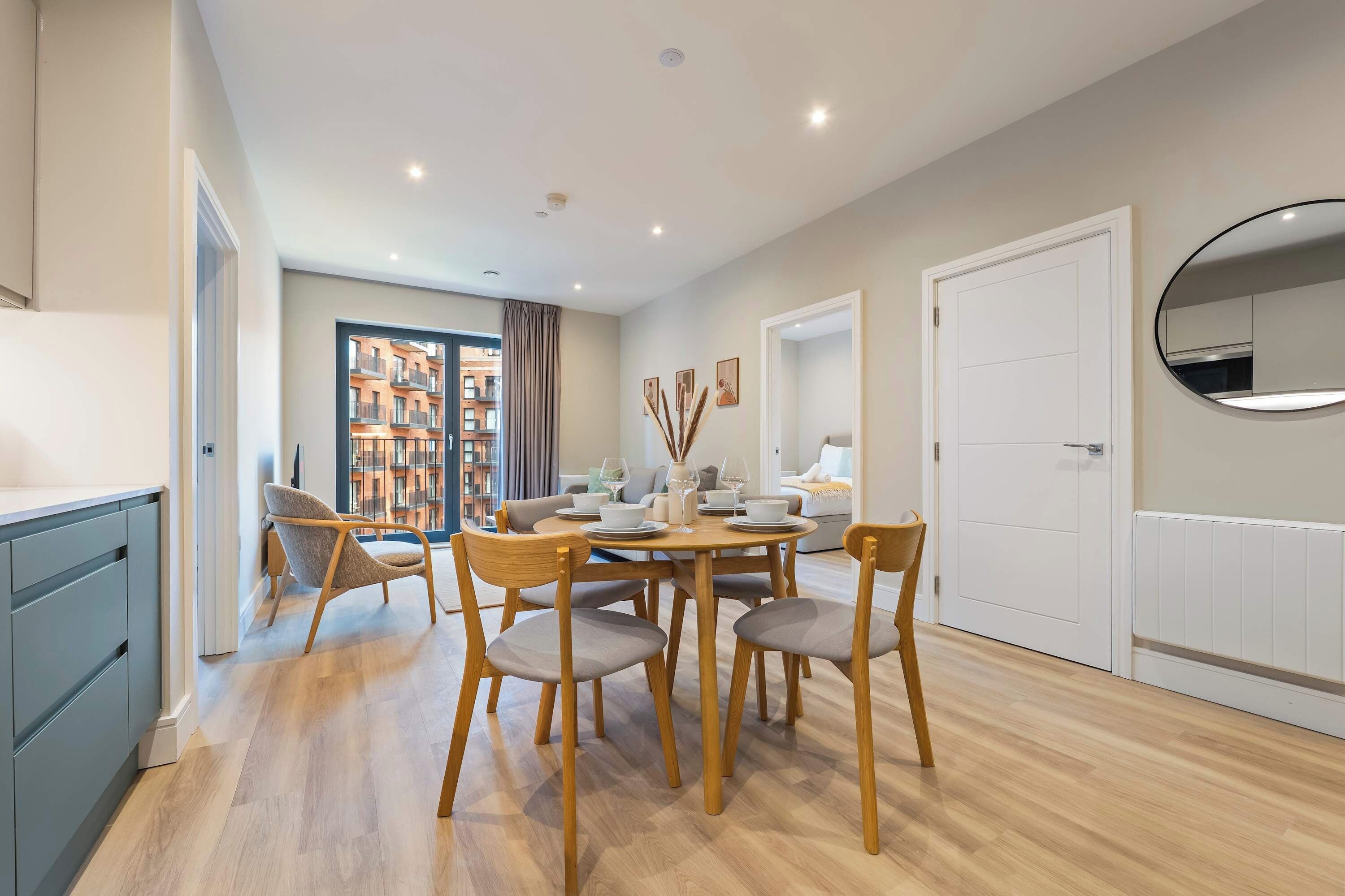 Un espace repas moderne avec une table ronde, des chaises en bois et une vue sur un immeuble par une grande fenêtre.
