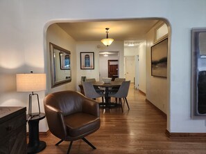 Un couloir mène à une salle à manger où se trouvent une table et des chaises, un fauteuil en cuir, une lampe et des tableaux encadrés accrochés aux murs.