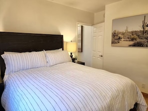 Ein Bett mit gestreifter Bettwäsche, ein Nachttisch mit Lampe, eine Tür und ein an der Wand hängendes Bild von einer Wüstenlandschaft.