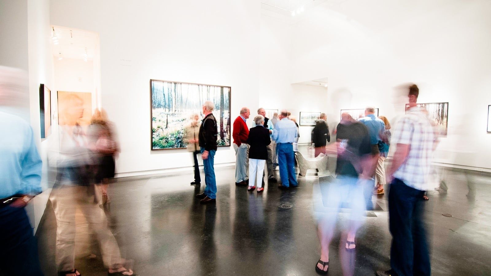 Una galería amplia con piso pulido, pinturas grandes en las paredes y un grupo de personas observando las obras de arte.