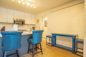 Una cocina moderna con armarios blancos, un microondas de acero inoxidable y una estufa a gas. Hay dos taburetes azules y una mesa pequeña azul.