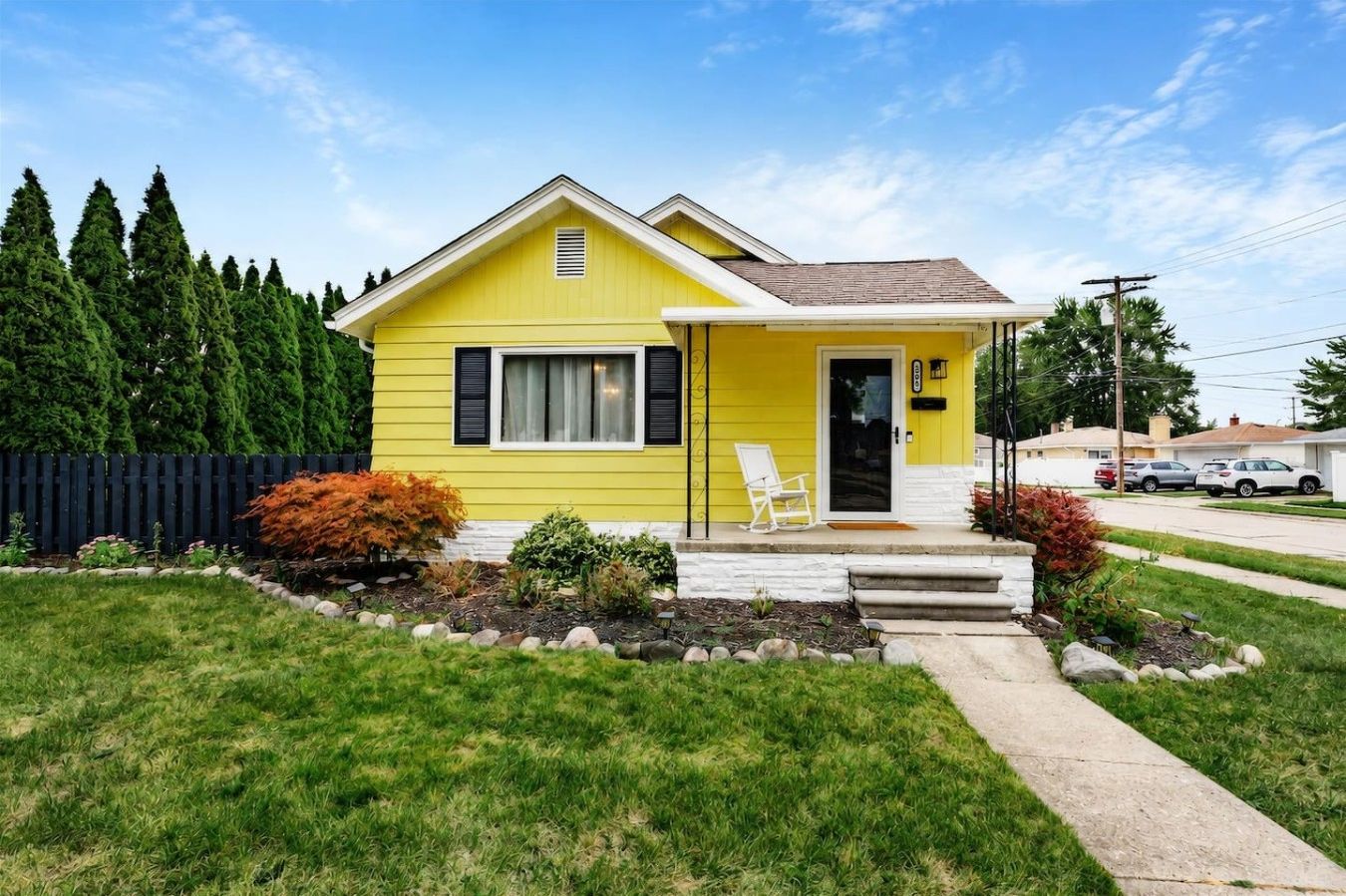 A yellow single-story house with a front porch, a small garden, and a black fence.