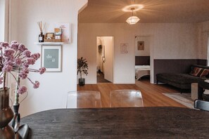 Una sala de estar con un sofá oscuro, una mesa de comedor de madera con sillas y un jarrón con flores. Hay un cuadro en la pared y una lámpara colgante.