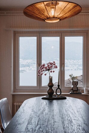 A dining table with a vase of flowers and a candle, set against a window with a view of a cityscape. - Casa 2 – Modern Vacation Rental on Lake Maggiore (Vairano)