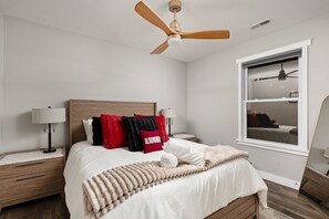 A bedroom with a bed, two bedside tables, a ceiling fan, a window with a view, and a mirror. - Hoosier Hideaway | King Bed, Beer Pong, Shotski (Bloomington)