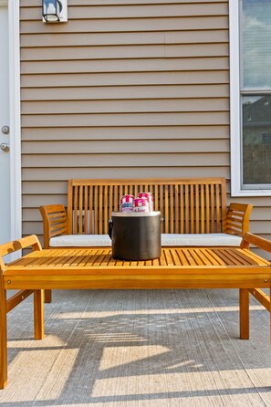 A wooden patio table with a metal bucket containing cans, positioned in front of a wooden bench with a slatted backrest. - Hoosier Hideaway | King Bed, Beer Pong, Shotski (Bloomington)