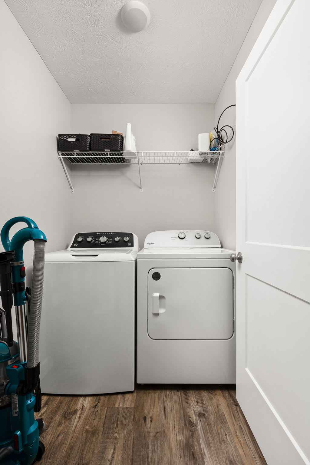A laundry room with two washing machines, a vacuum cleaner, and a shelf with items.