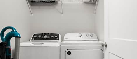 A laundry room with two washing machines, a vacuum cleaner, and a shelf with items.