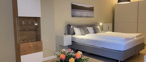 A modern bedroom with a bed, bedside tables, a lamp, a wall-mounted artwork, and a vase with flowers.