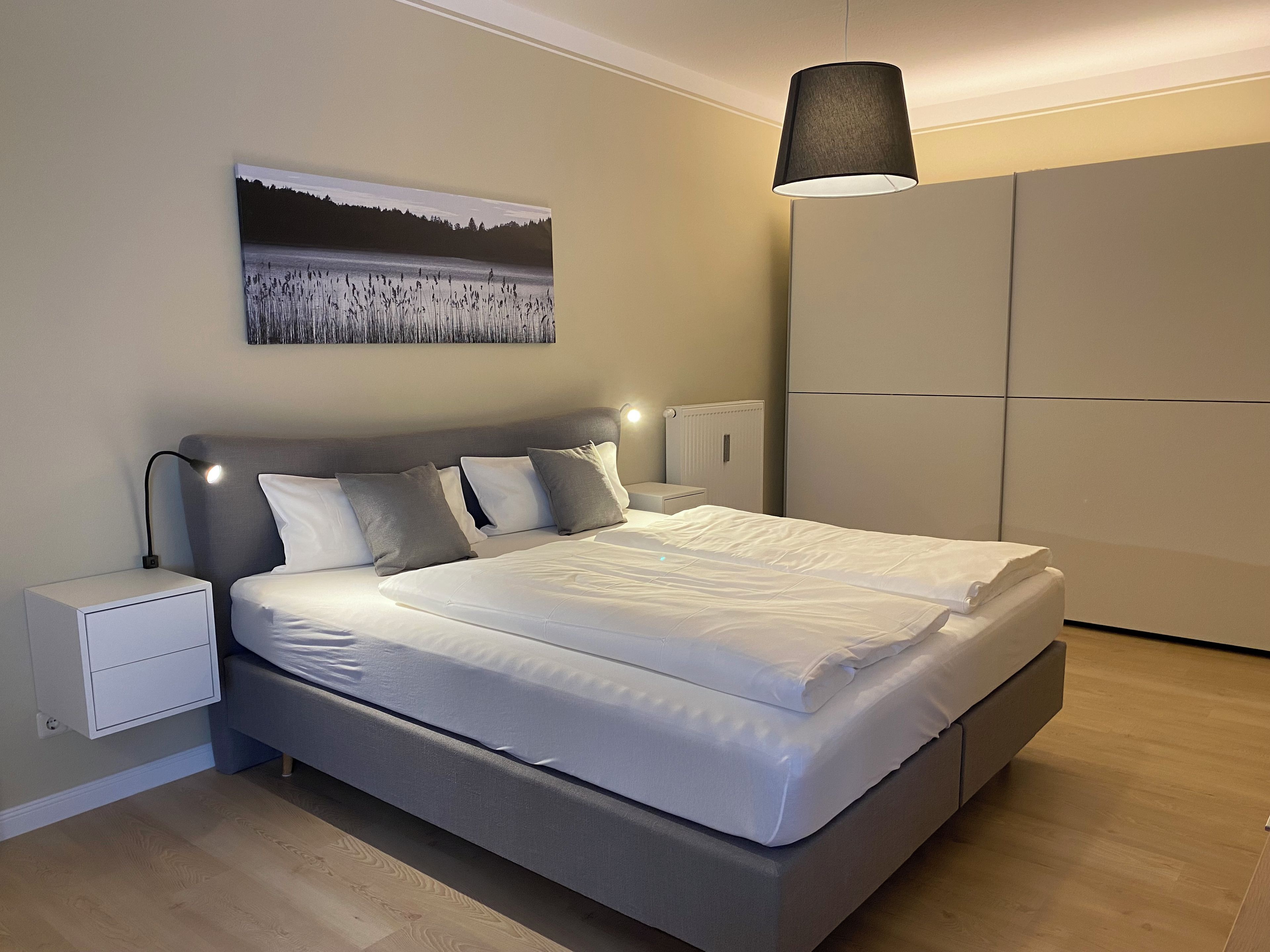 A modern bedroom with a large bed, bedside tables, a hanging lamp, and a painting on the wall.