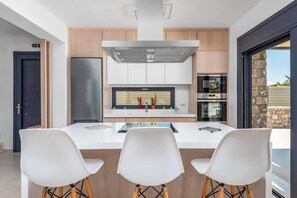 A modern kitchen with a central island, stainless steel appliances, and a view to the outside. - Villa Artemis Lindian Palm by Villa Plus (Lardos)