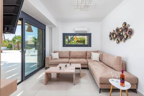 Een moderne woonkamer met een beige hoekbank, een houten salontafel en een klein wit bijzettafeltje met een fles en glazen.