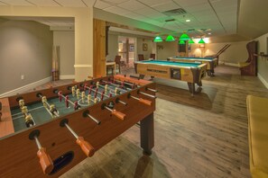 Una habitación con mesa de fútbol de mesa y mesa de billar, suelo de madera y lámparas colgantes verdes.