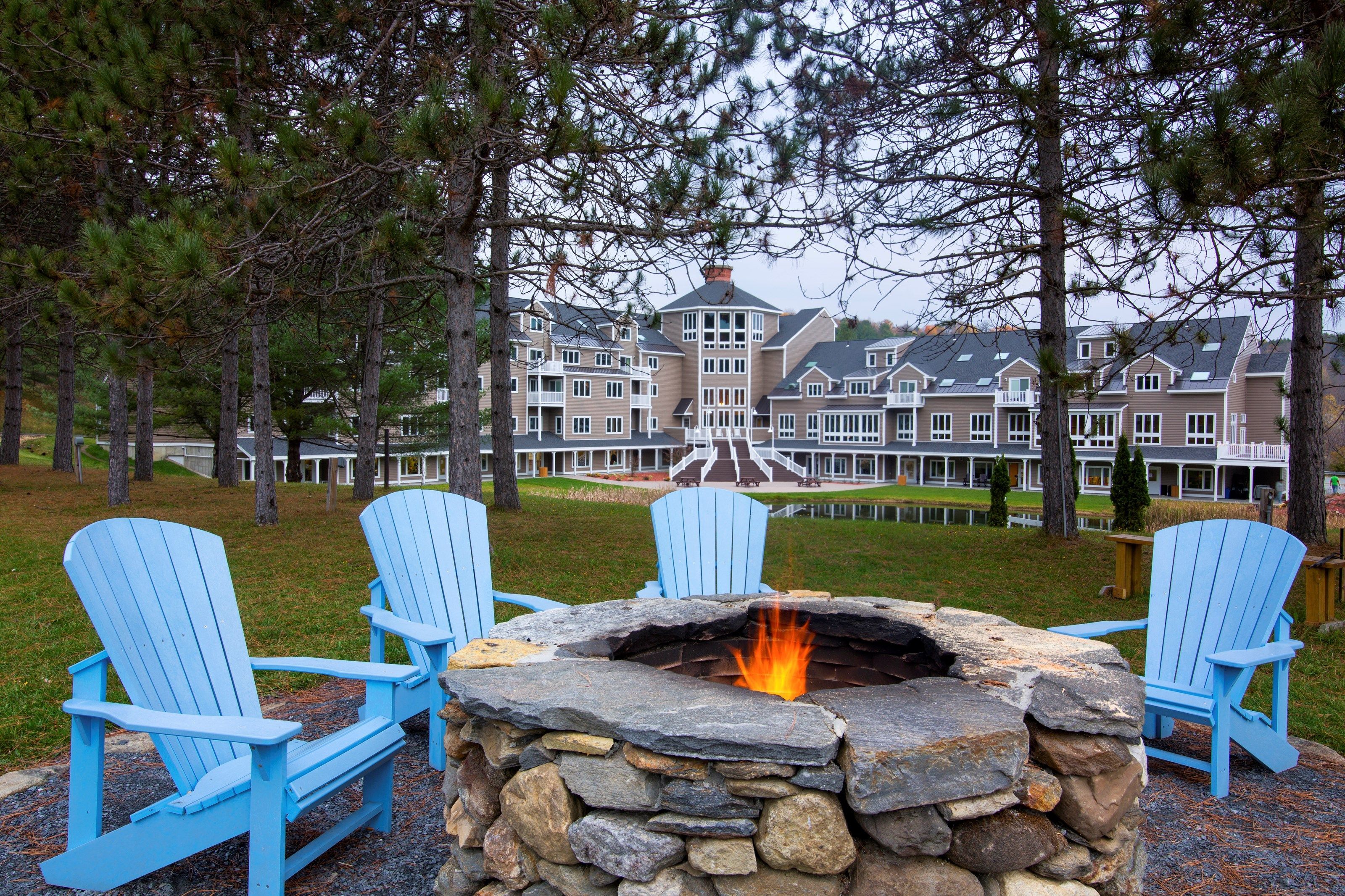 Een vuurkorf van steen met blauwe Adirondack-stoelen ervoor, omringd door bomen.
