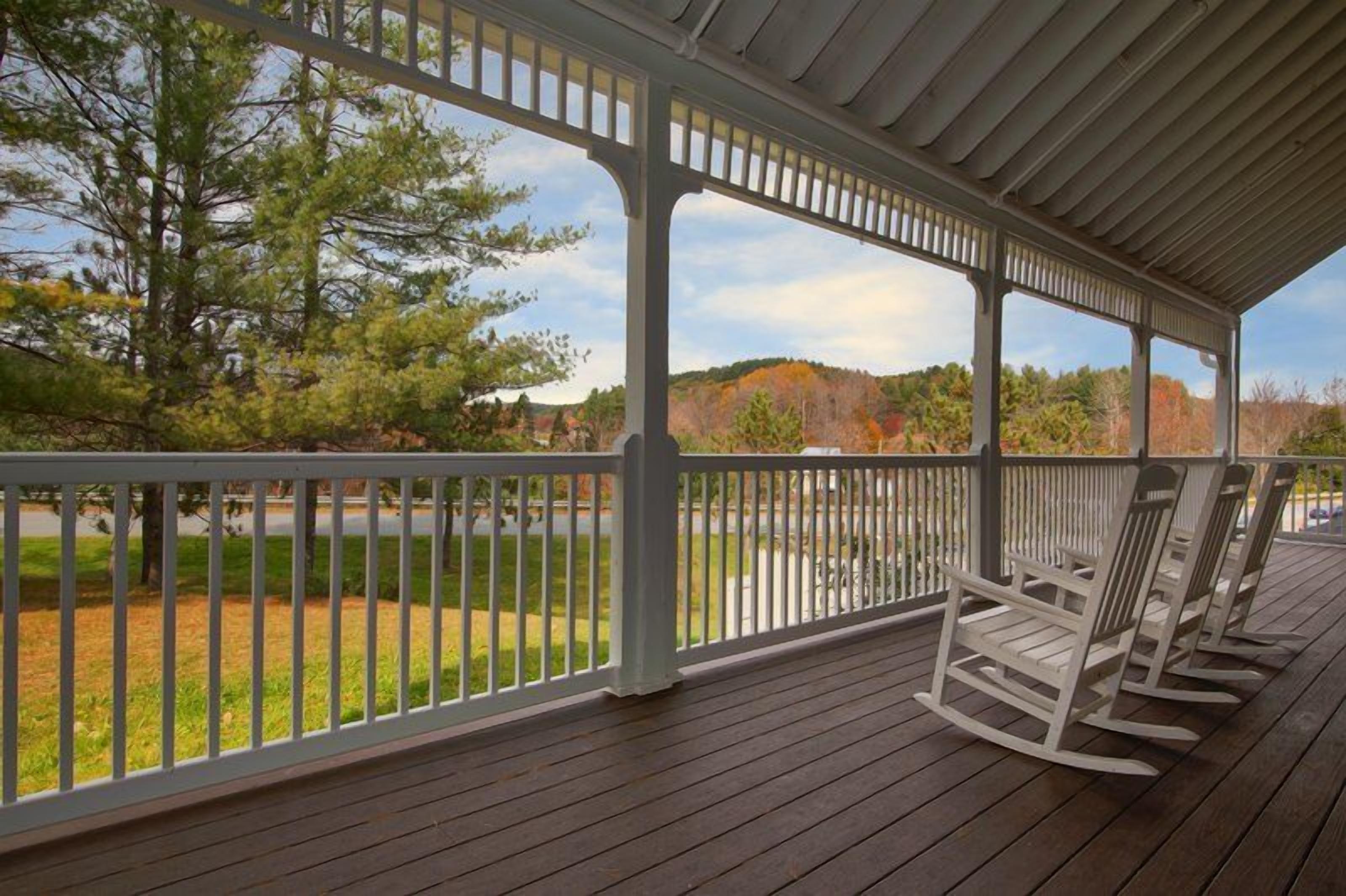 A covered porch with white rocking chairs, a wooden deck, and a view of trees and hills.