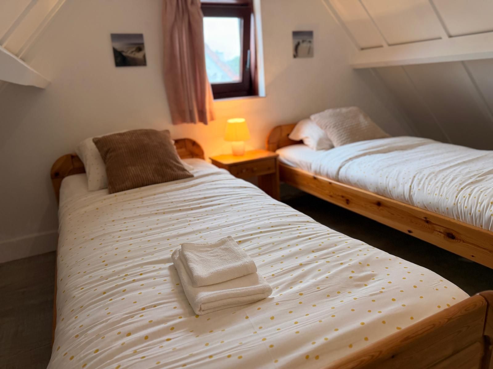 Quarto com duas camas de solteiro, mesa de cabeceira, luminária e janela com cortinas.