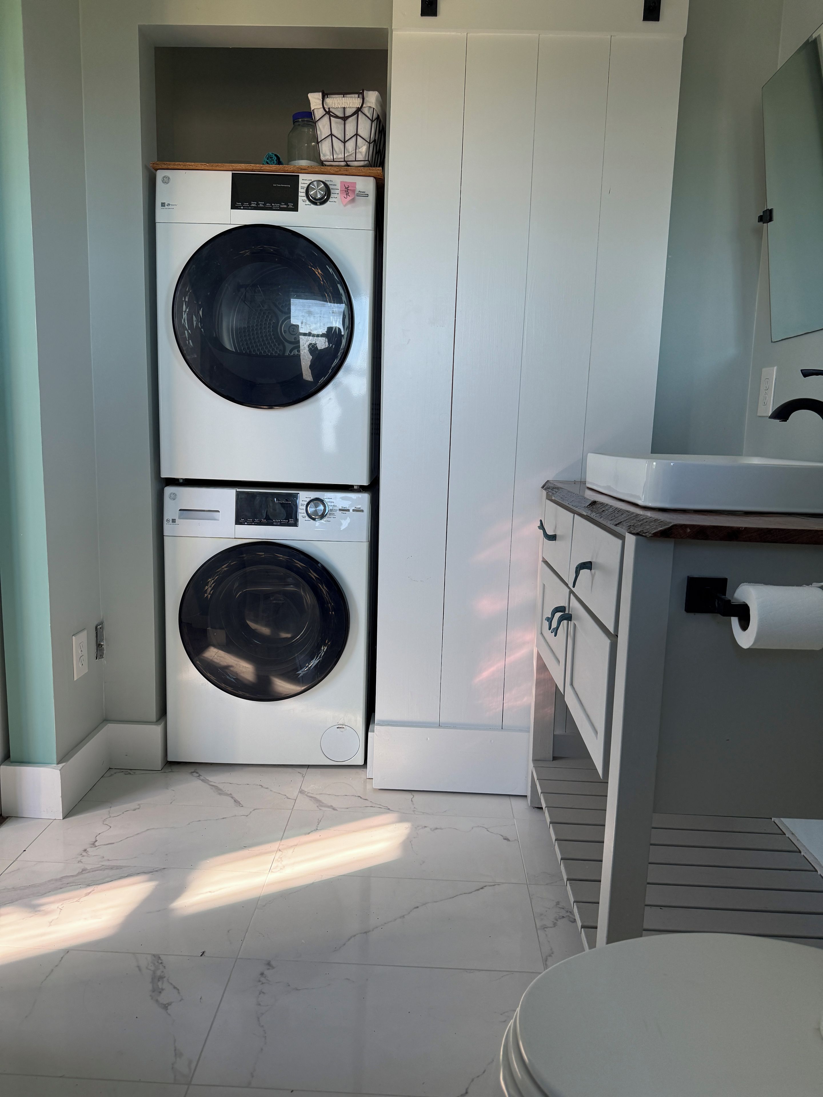A compact laundry room with a front-loading washer and dryer, a small shelf with containers, and a bathroom with a toilet paper holder.