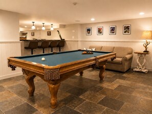 Una mesa de billar con taco, un sofá y una lámpara en una habitación con piso de baldosas y una zona de bar al fondo.