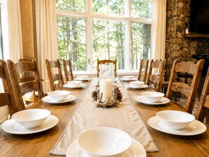 Una mesa de comedor lista para una comida, con platos y tazones blancos, un centro de mesa con vela y vistas a árboles a través de ventanas amplias.