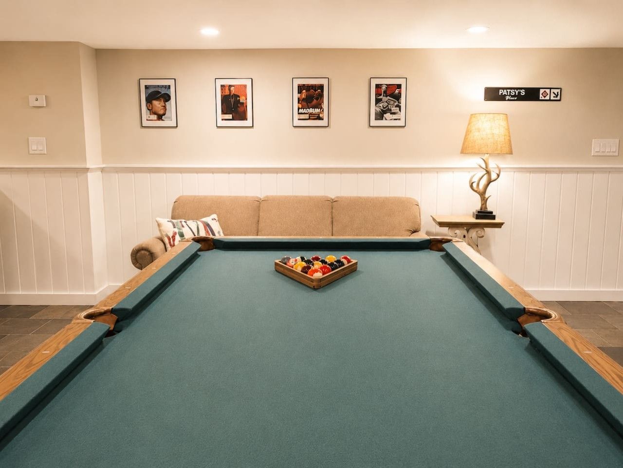 A pool table with a cue ball and a rack of billiard balls, a sofa, a lamp, and framed pictures on the wall.