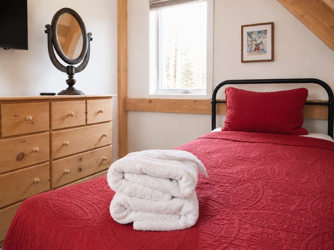 A bedroom with a bed, a red pillow, a wooden dresser, a mirror, and a window.