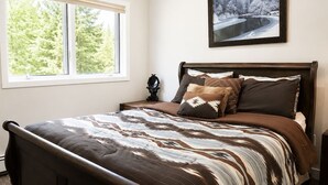Een slaapkamer met een groot bed, een nachtkastje, een raam met uitzicht op bomen en een lijst met een afbeelding aan de muur.