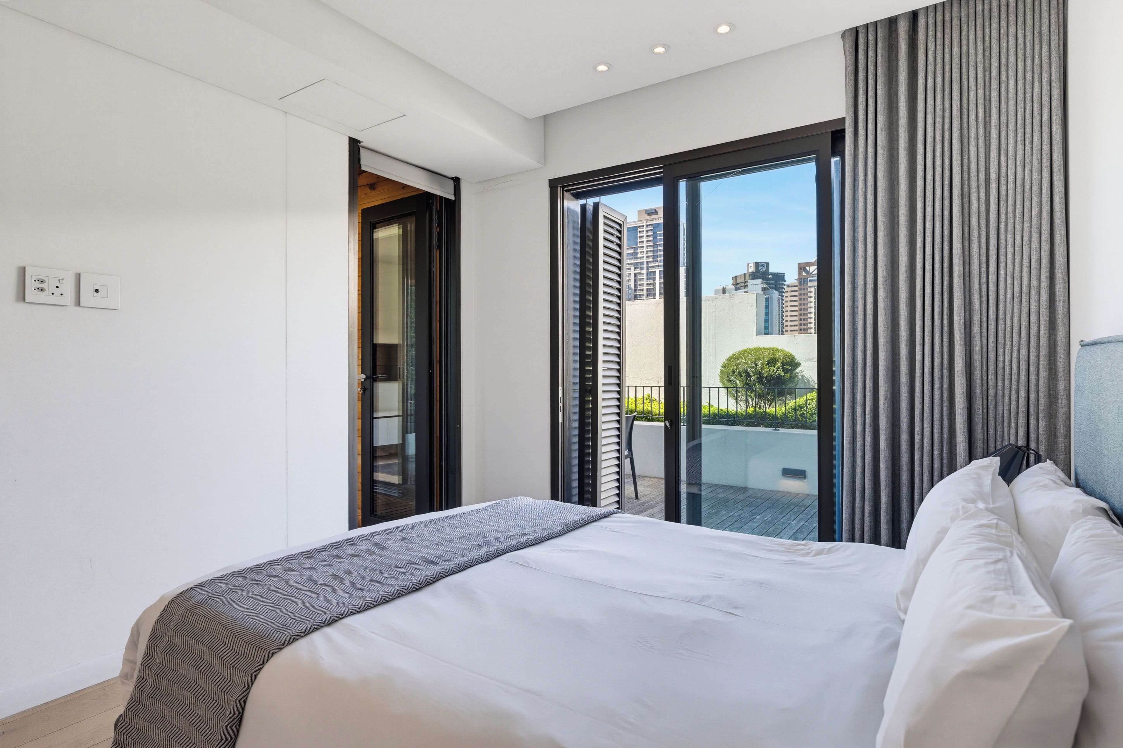 A modern hotel room with a large bed, a sliding glass door to a balcony, and a city view.