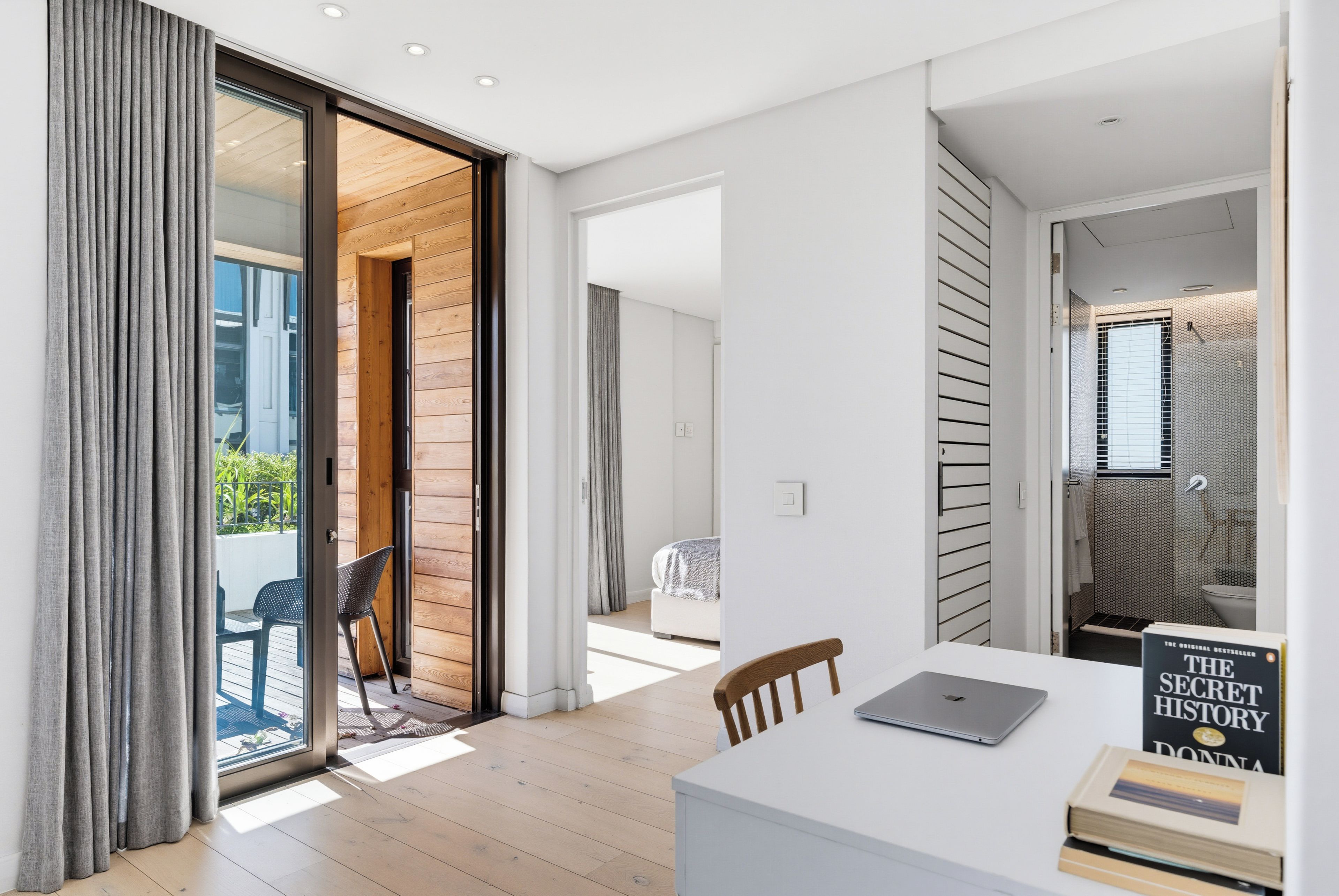 Een modern interieur met een schuifglazen deur naar een balkon, een wit bureau met een laptop en een boek getiteld 'The Secret History' van Donna Tartt.