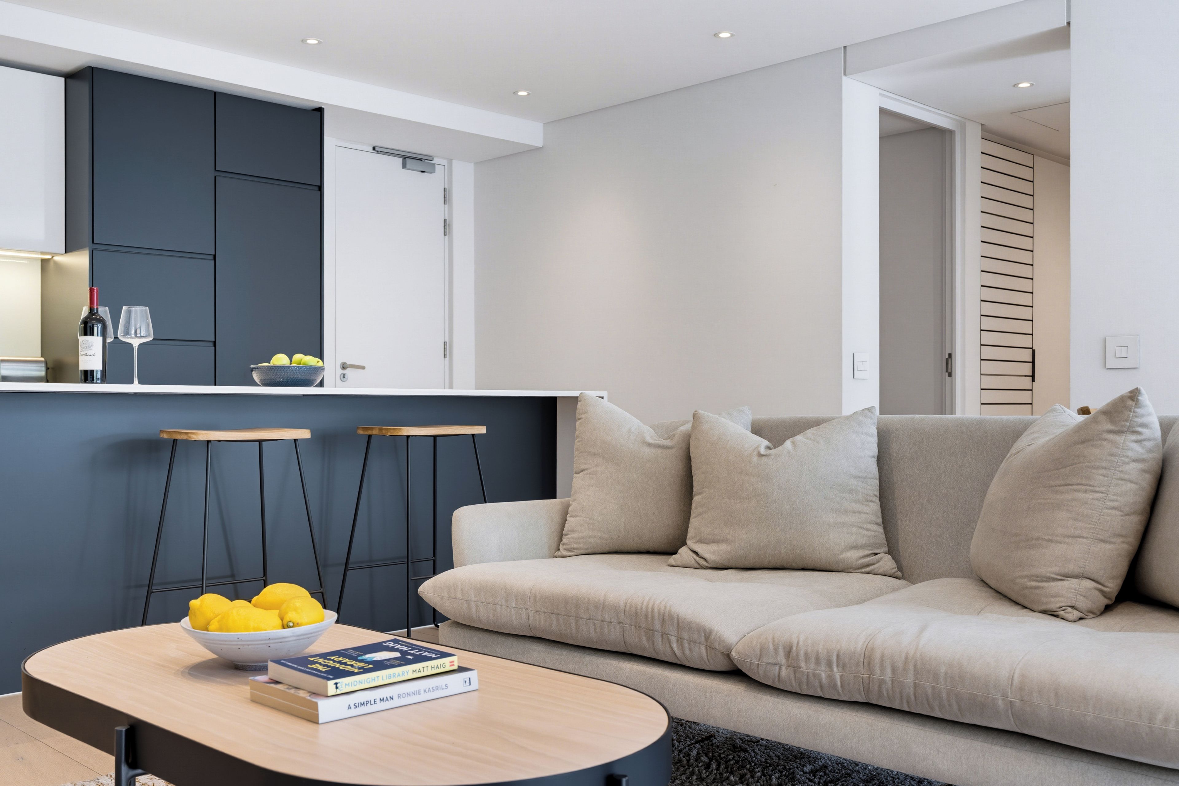 A modern living room with a sofa, a coffee table with books and lemons, and a kitchen area with cabinets and a wine bottle.