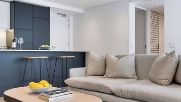 A modern living room with a sofa, a coffee table with books and lemons, and a kitchen area with cabinets and a wine bottle.