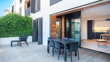 A modern outdoor dining area with a wooden deck, black furniture, and a glass door leading to a kitchen.