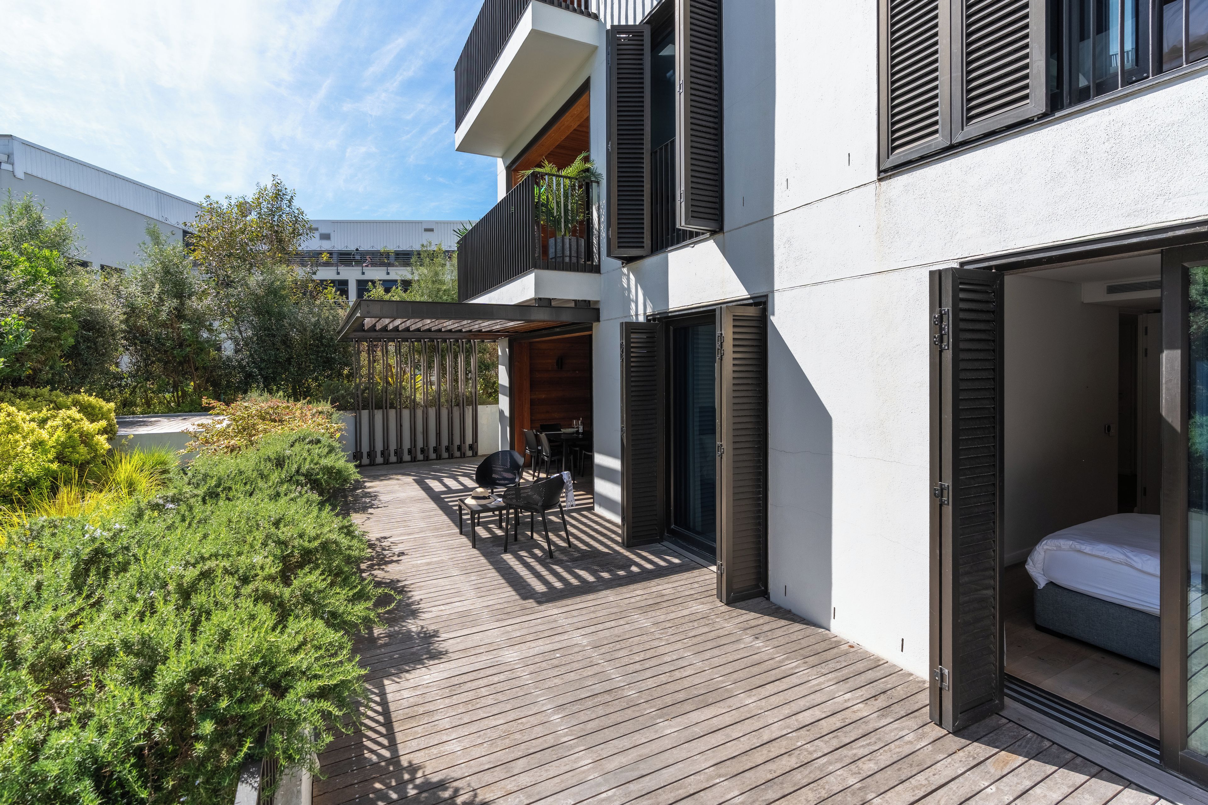 Une terrasse extérieure moderne, dotée d’un pont en bois, d’un espace salon avec des chaises et une table, et d’une porte vitrée donnant accès à une chambre.