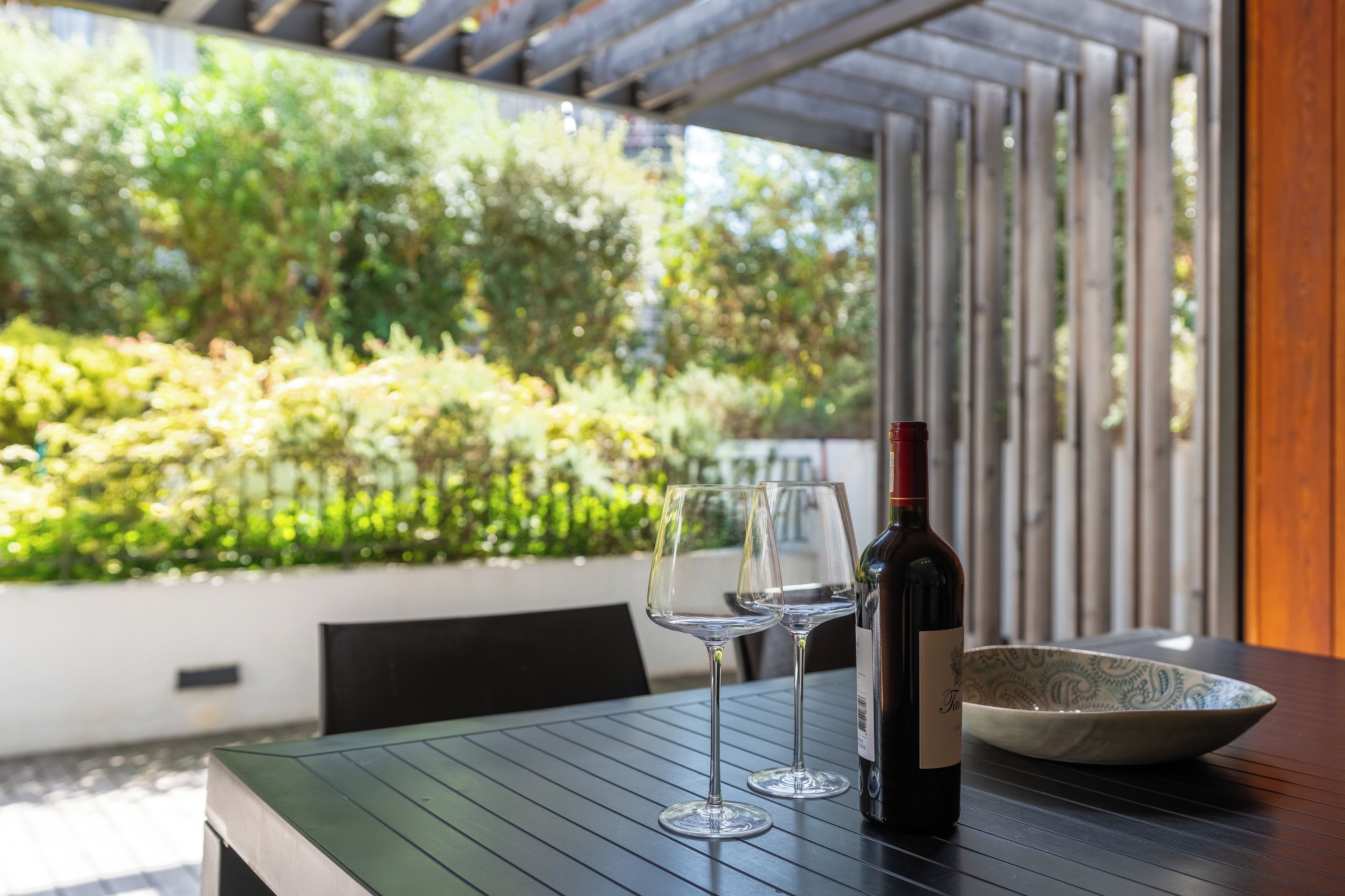 Ein Tisch mit einer Weinflasche und zwei Gläsern, einer Schüssel sowie Blick auf einen grünen Garten durch ein Fenster.