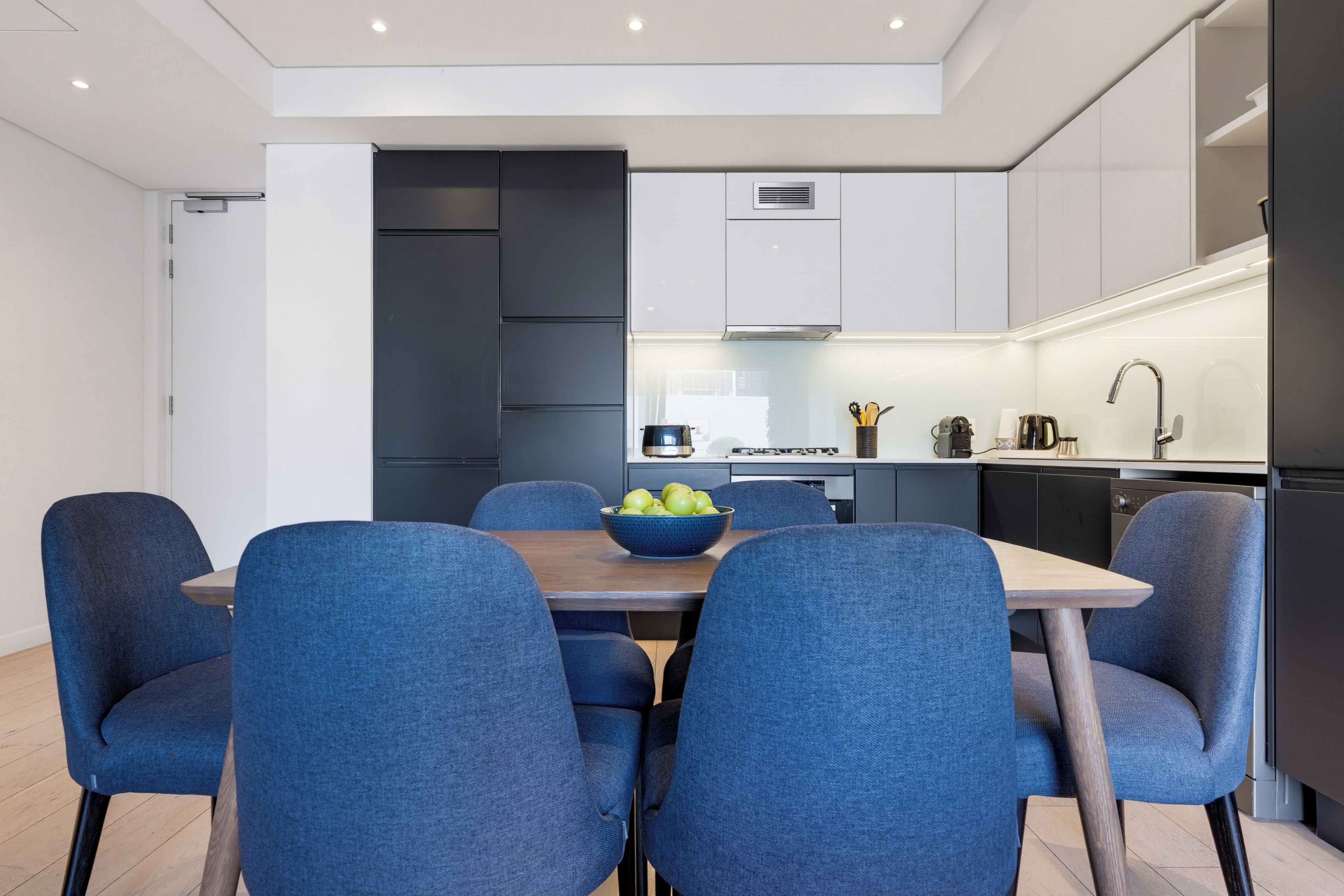 Une cuisine moderne avec des meubles blancs, une table à manger avec des chaises bleues et un saladier de fruits posé sur la table.