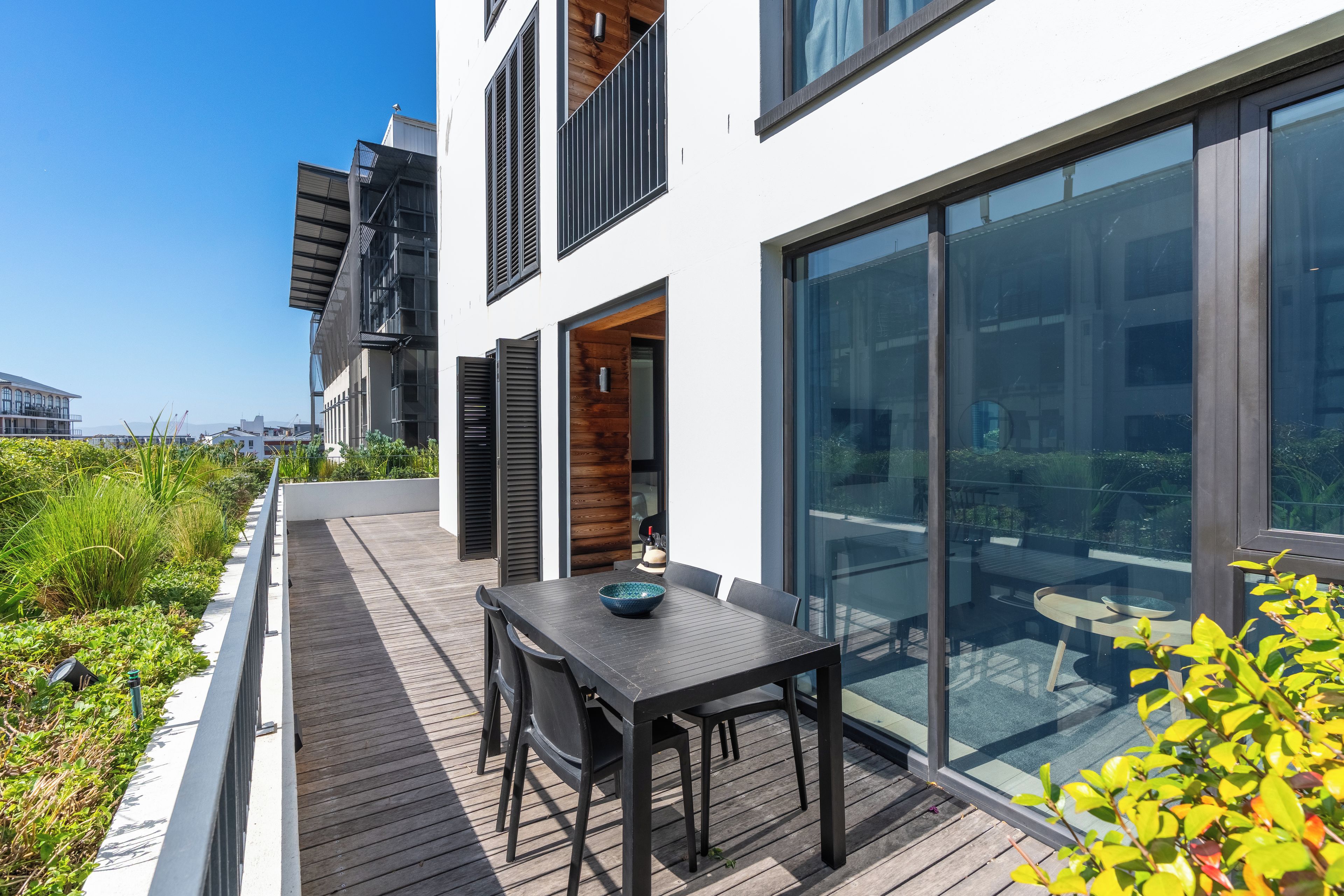 A modern outdoor patio with a table and chairs, a wooden deck, and a glass door leading to a balcony.