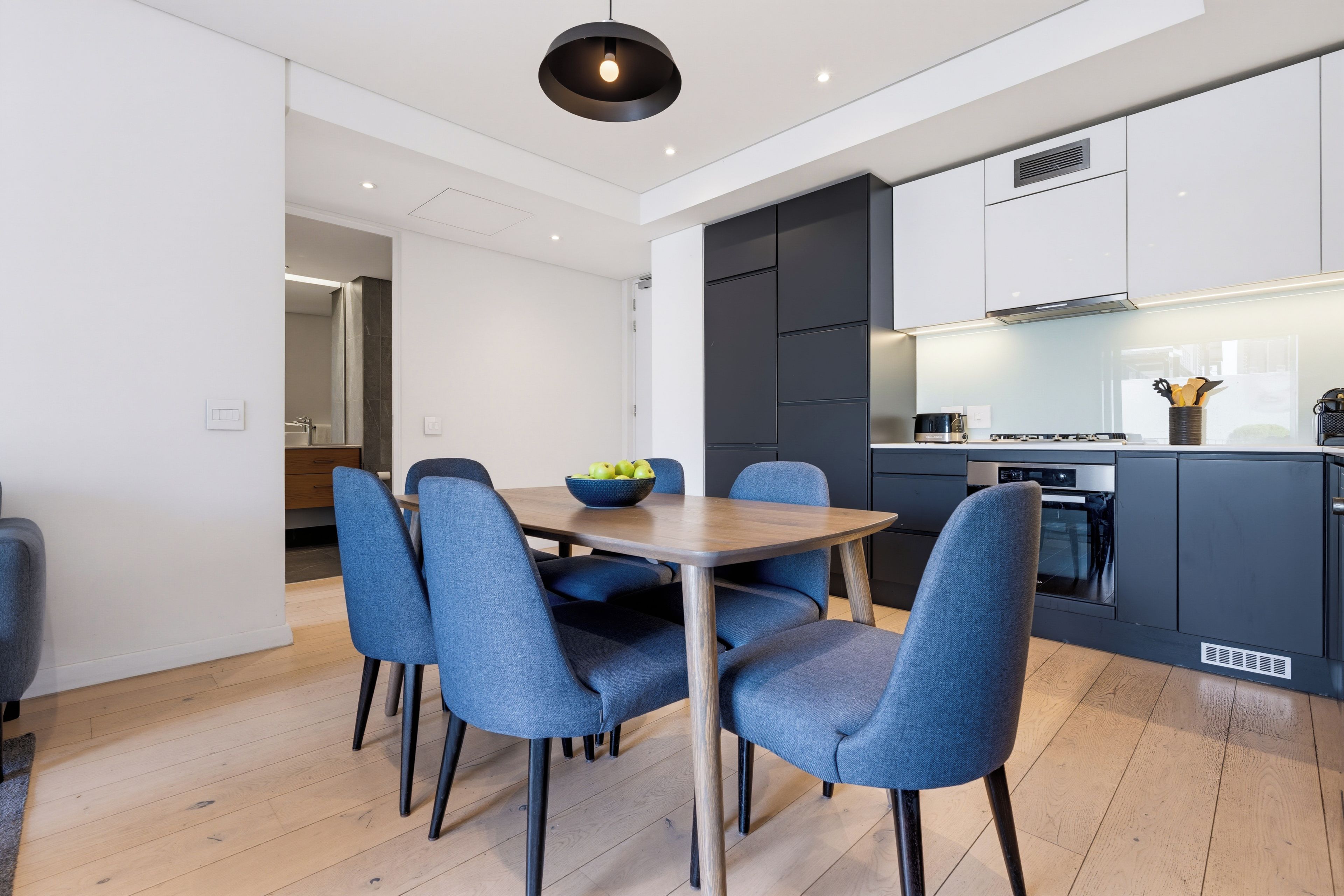 Una cocina moderna con armarios oscuros, horno empotrado y zona de comedor con mesa de madera y sillas tapizadas en azul.