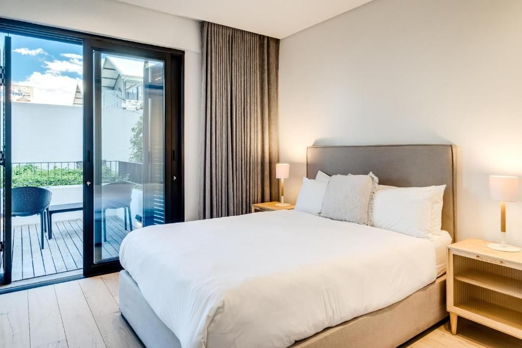 A hotel room with a large bed, a nightstand, a lamp, and a sliding glass door leading to an outdoor area with a chair.