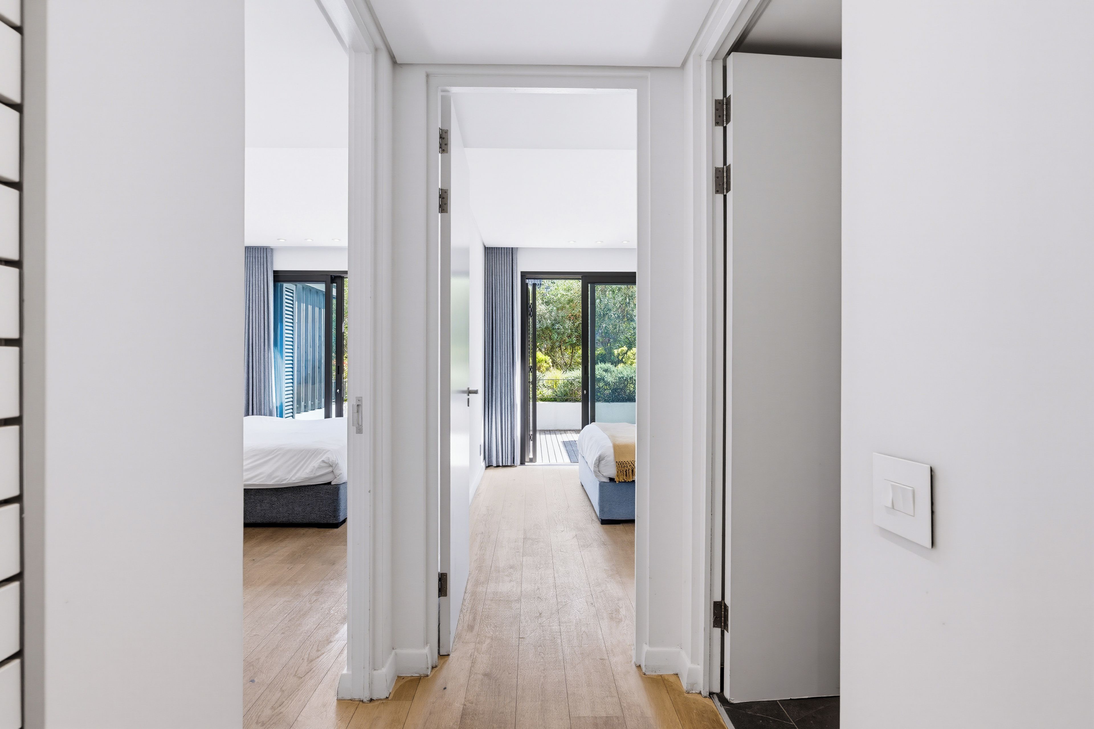 Un couloir mène à deux chambres dotées de grandes fenêtres, d’un plancher en bois et offrant une vue sur la verdure environnante.